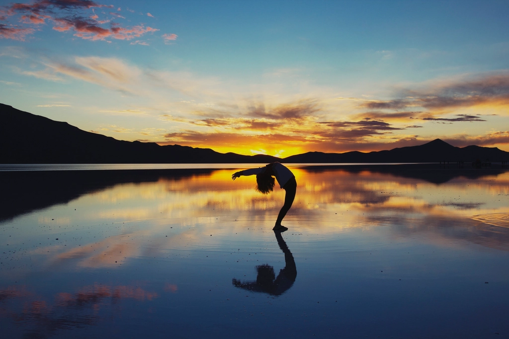 Silhouette of a person in a yoga backbend at sunset, reflected in still water